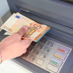 Dłoń z banknotami 50 euro wkłada pieniądze do bankomatu.