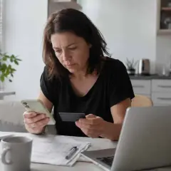 Zmartwiona kobieta sprawdza telefon i kartę płatniczą, podejrzewając nieuprawnione użycie. Obok laptop i dokumenty.