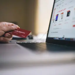 Dłoń trzyma czerwoną kartę, gotową do zapłaty online. Widok ekranu laptopa z ofertami sklepu internetowego pokazuje, jak placic karta.