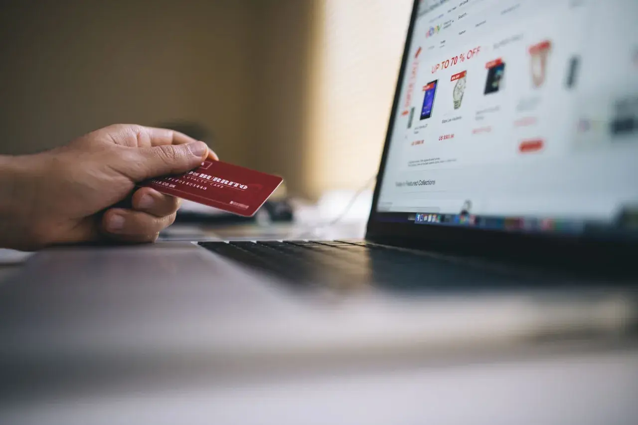 Dłoń trzyma czerwoną kartę, gotową do zapłaty online. Widok ekranu laptopa z ofertami sklepu internetowego pokazuje, jak placic karta.