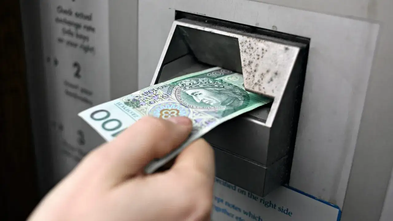 Ręka wkłada banknot 100 zł do bankomatu. W razie wojny czy państwo może zabrać pieniądze z konta?